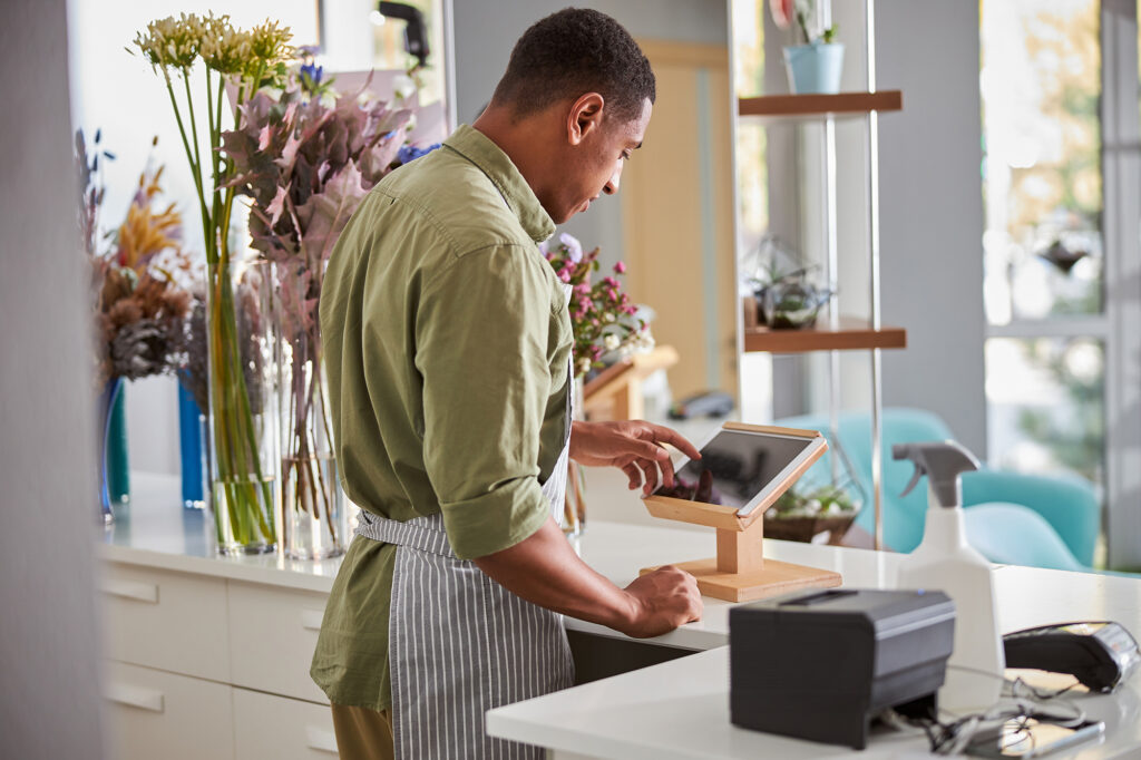 A black main in a flower shop, wearing a white apron and working on a point of sale system. He's wearing a green shirt.