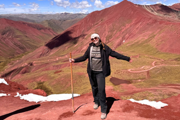 Young woman in dark, warm hiking clothes on a red mountain in Peru, with a walking stick.