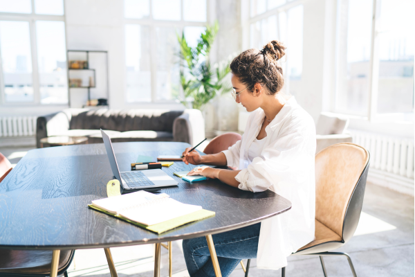 White woman with curly blond hair pulled into a bun, in a white shirt sitting at a table in an apartment, lots of white light, working on a laptop.